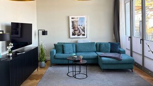 A modern living room with a sectional sofa, a flat-screen TV, a small round table, and a large window with curtains. - Luxury Apartment "Strandperle" – First Row | Private Sauna | Family & Dog | Ocean View (Binz)