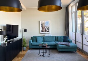 Ein modernes Wohnzimmer mit einem Ecksofa, einem Flachbildfernseher, einem kleinen runden Tisch und einem großen Fenster mit Vorhängen.