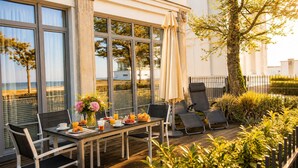 A sunlit terrace with a table set for breakfast, overlooking a beach. - Luxury Apartment "Strandperle" – First Row | Private Sauna | Family & Dog | Ocean View (Binz)