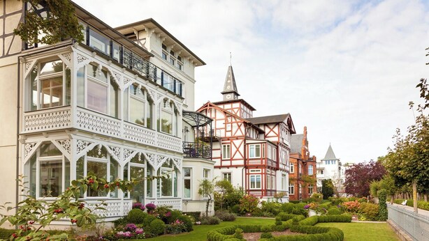 A historic building with a balcony, a garden with hedges, and a church in the background. - Luxury Apartment "Strandperle" – First Row | Private Sauna | Family & Dog | Ocean View (Binz)