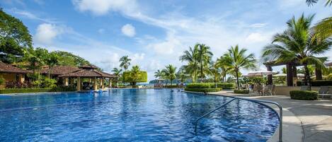 A large outdoor swimming pool with a clear blue sky, surrounded by palm trees and lounge chairs.