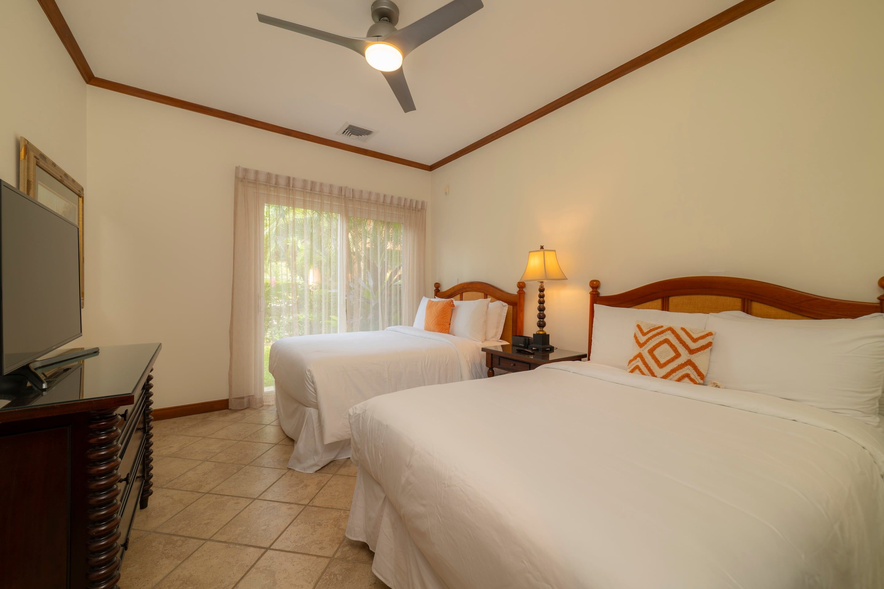 A hotel room with two beds, a ceiling fan, a desk with a television, and a window with curtains.