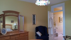 Une chambre à coucher avec une commode en bois, un lit recouvert d’une couvre-lit à motifs floraux, un fauteuil sombre, un lustre en vitrail et une salle de bain visible à travers une porte ouverte.