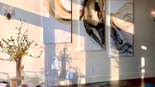 Une pièce dans laquelle on trouve deux grands tableaux accrochés au mur, un vase contenant des fleurs séchées et une table en verre surmontée de beaux verres transparents.
