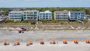 Ein Strandhotel mit mehreren Gebäuden, einem Sandstrand mit Sonnenschirmen und einem klaren Himmel.