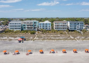 Un hôtel en bord de mer, composé de plusieurs bâtiments à étages, avec une plage de sable dotée de parasols et offrant une vue dégagée sur l’océan.