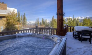 A hot tub with a snowy deck, wooden chairs, and a mountain view.