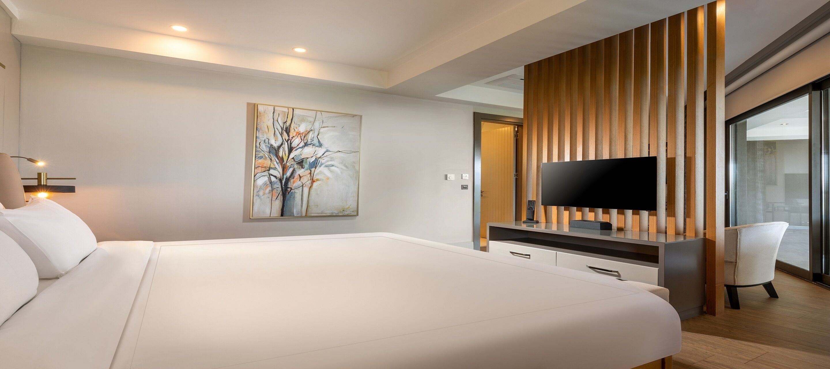 A modern hotel room with a large bed, a flat-screen TV, a wooden panel wall, and a white chair.