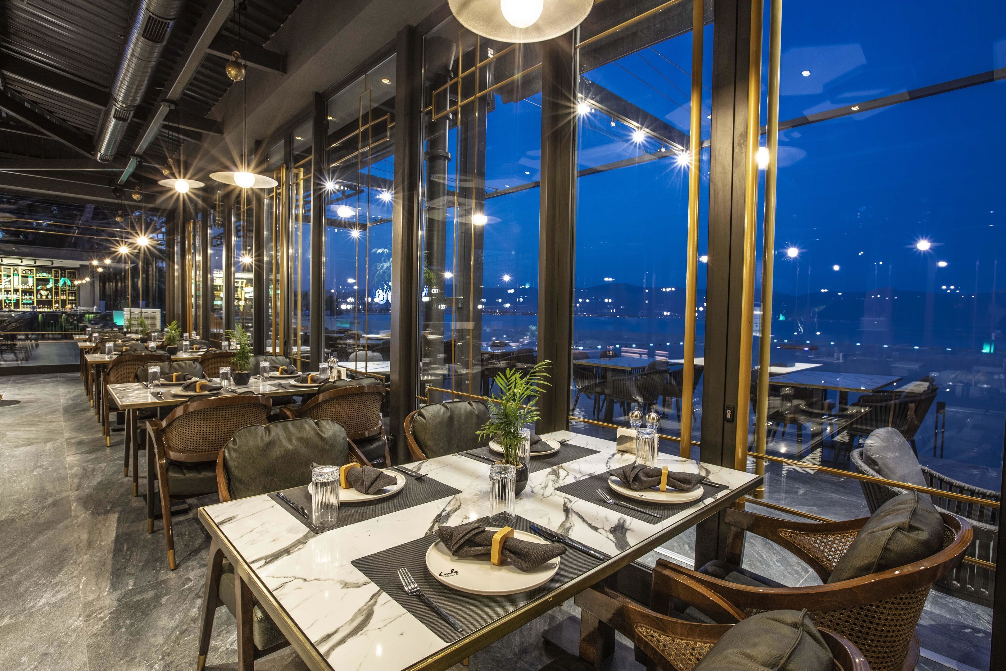 A modern restaurant with marble-topped tables, wicker chairs, and a city view at night.