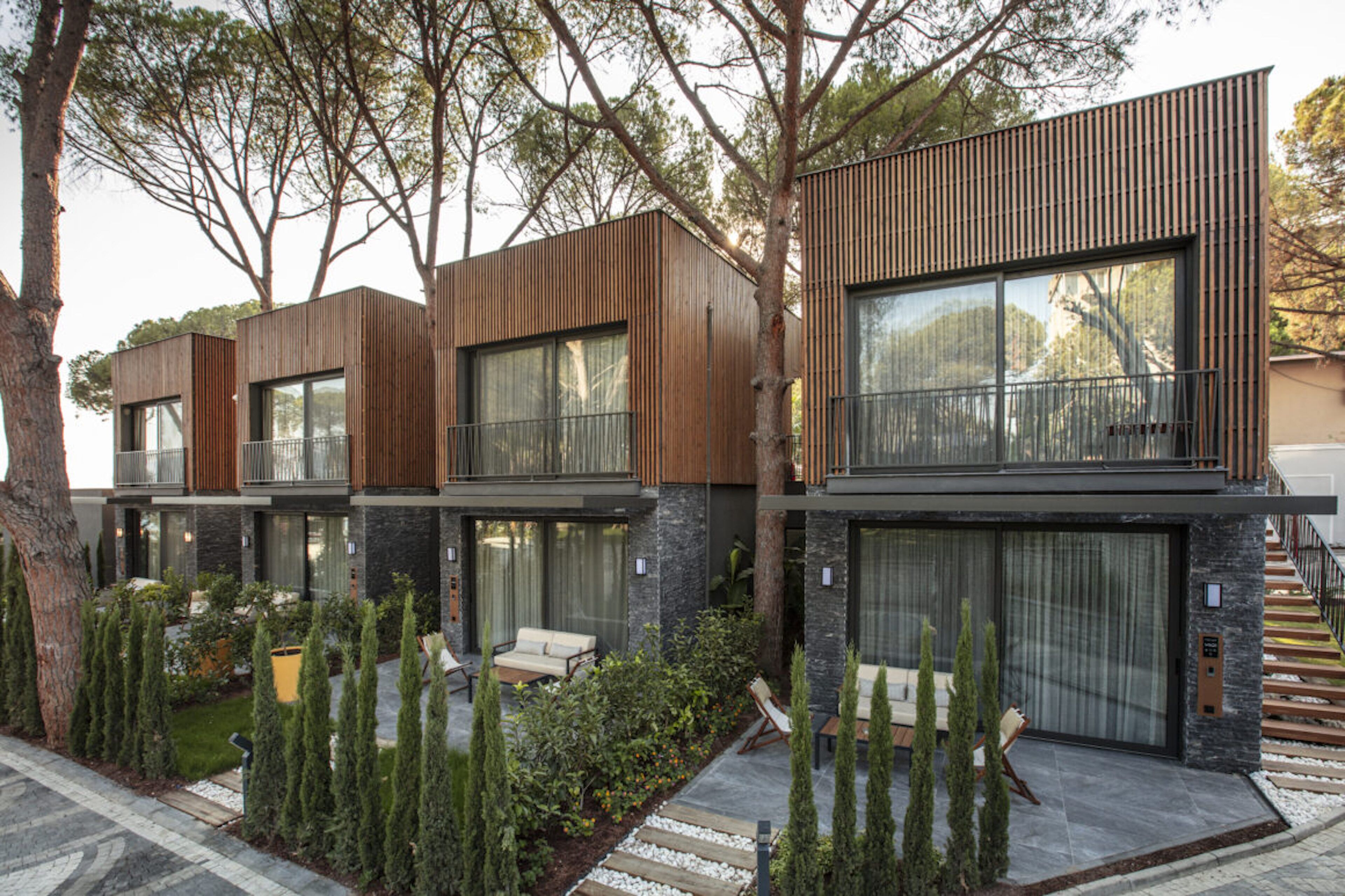 Un moderno edificio residencial de varios pisos, con amplios ventanales, un balcón y un jardín ajardinado con cipreses y un sendero.