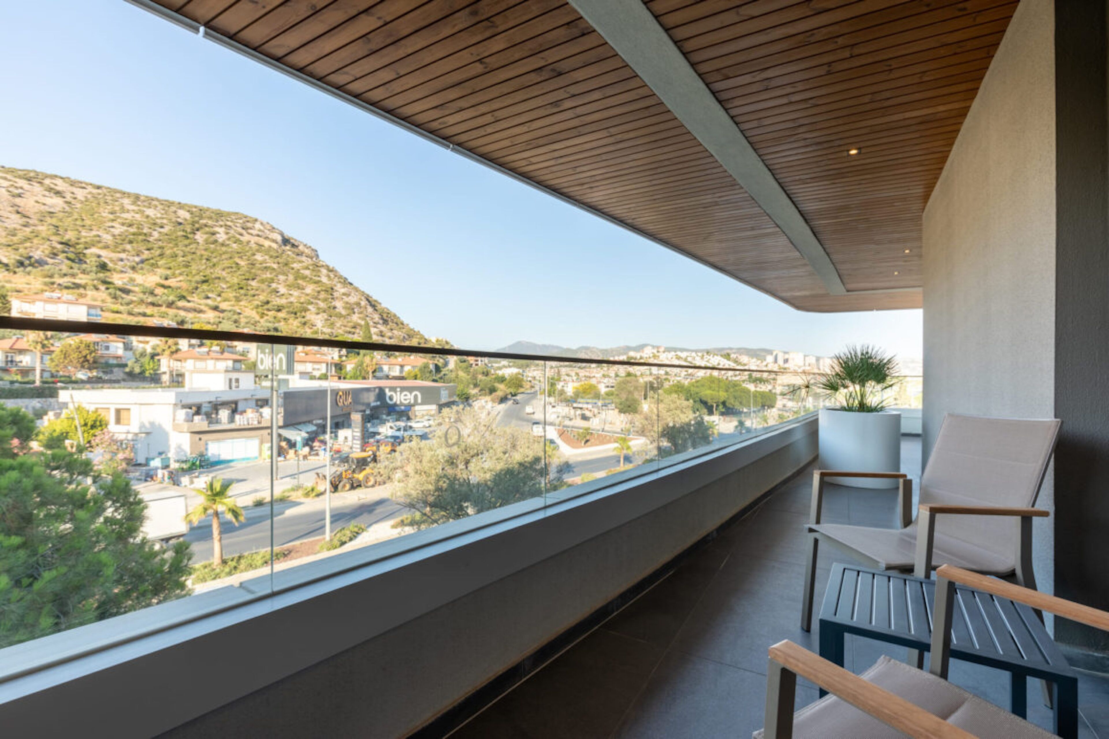 A balcony with a view of a hilly landscape and buildings. There are two chairs and a small table on the balcony.
