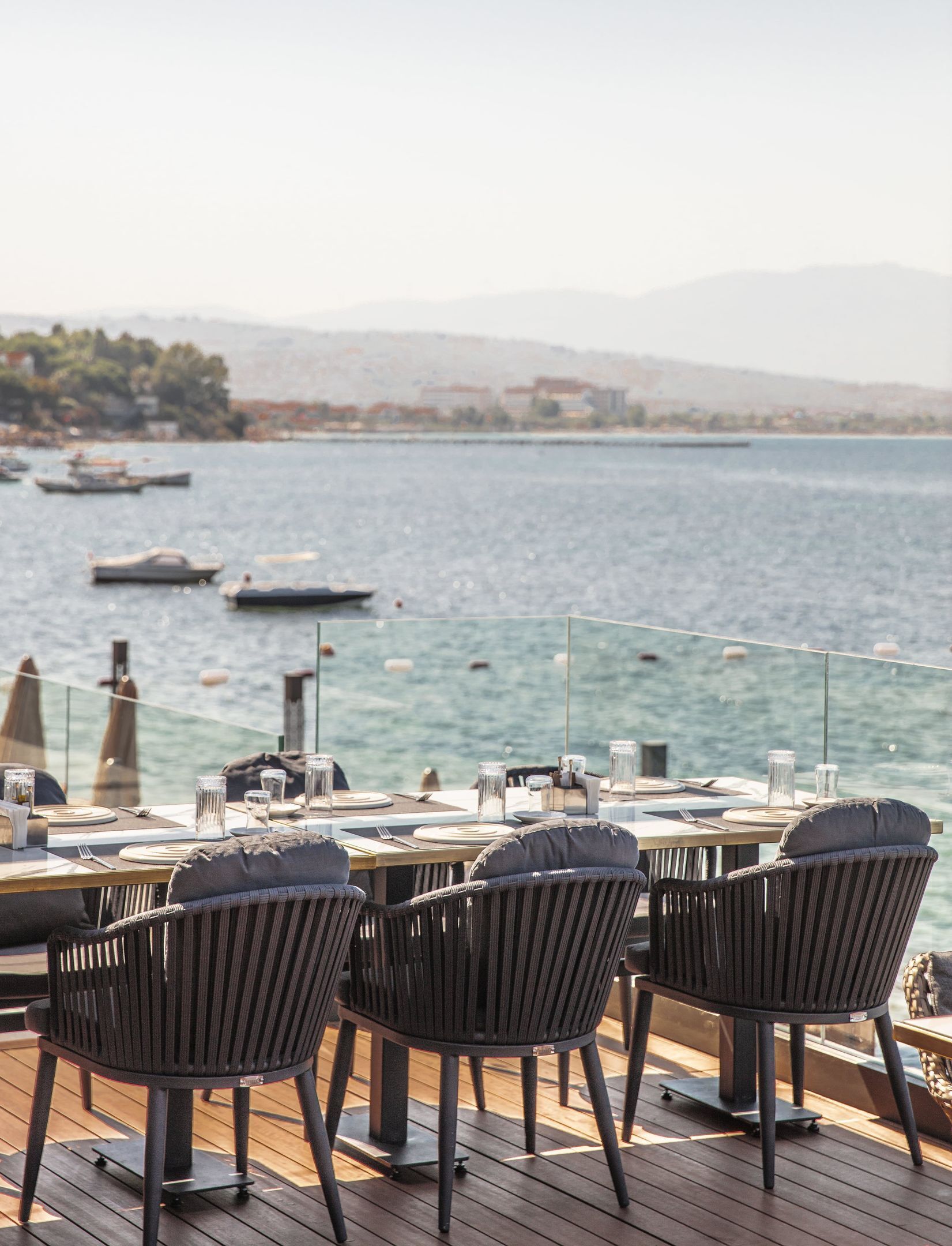 An outdoor dining area with tables and chairs, overlooking a body of water with boats and a distant shoreline.