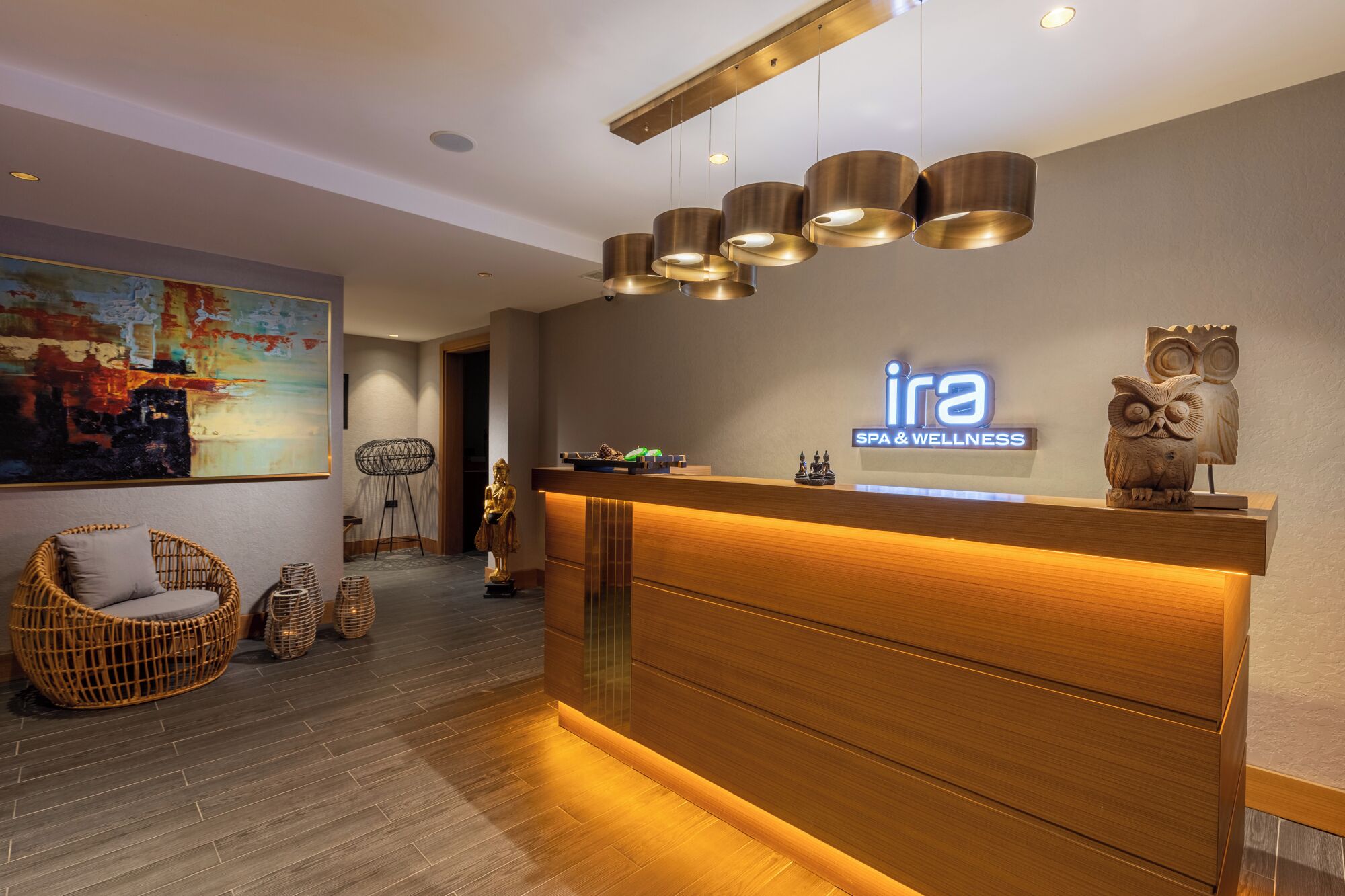 Ein Empfangsbereich mit einer Rezeption, an der das Schild 'ira SPA & WELLNESS' angebracht ist, Holzmöbel, Korbstühle und einem Gemälde an der Wand.