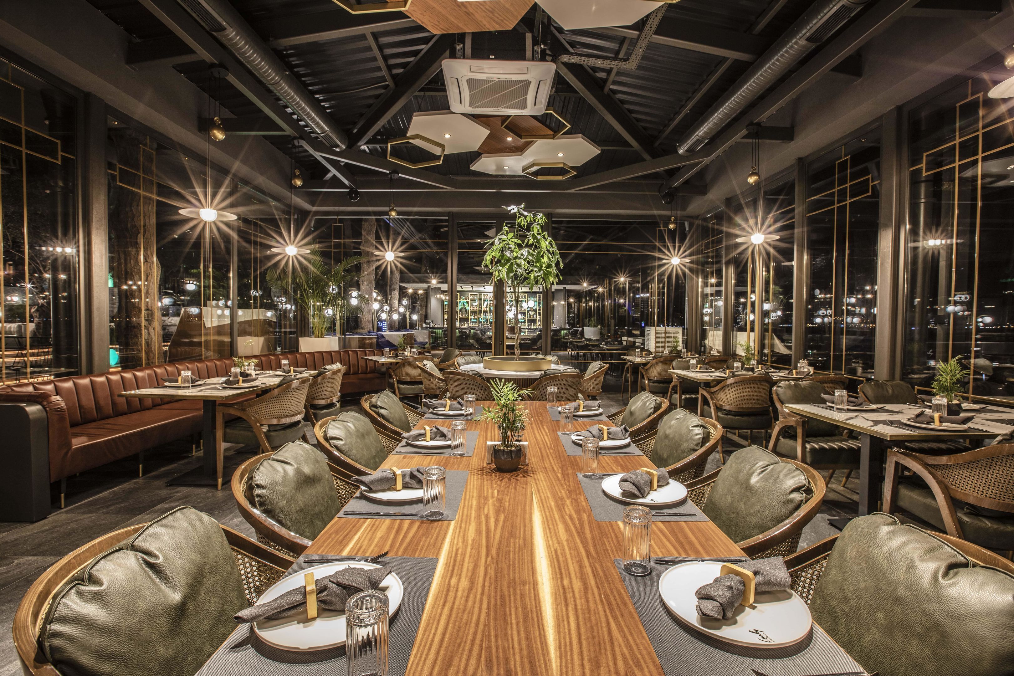 A modern restaurant with a long wooden table, leather chairs, and a central plant.