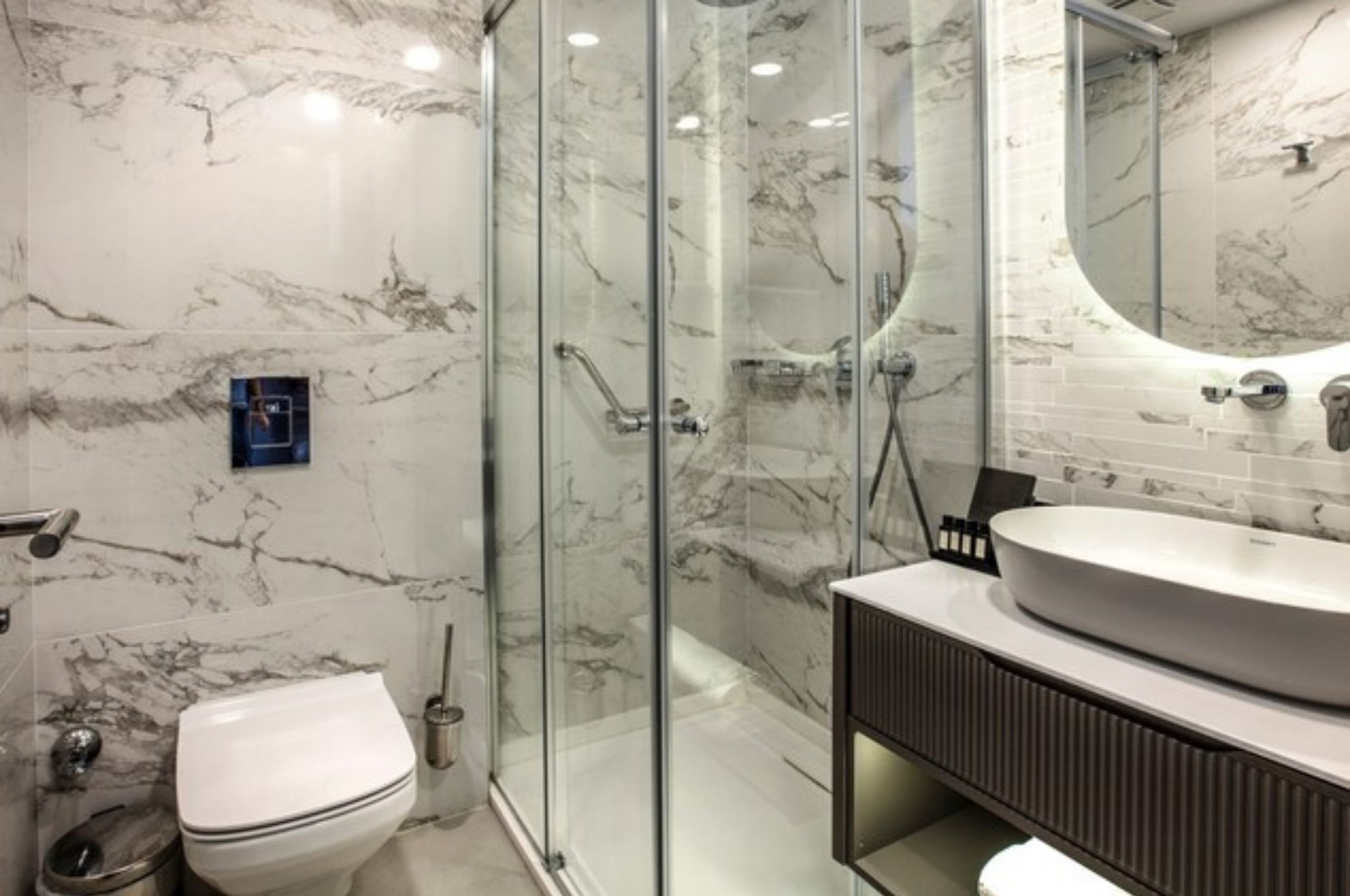 Un baño moderno con ducha de vidrio, inodoro blanco y lavamanos redondo sobre encimera oscura.