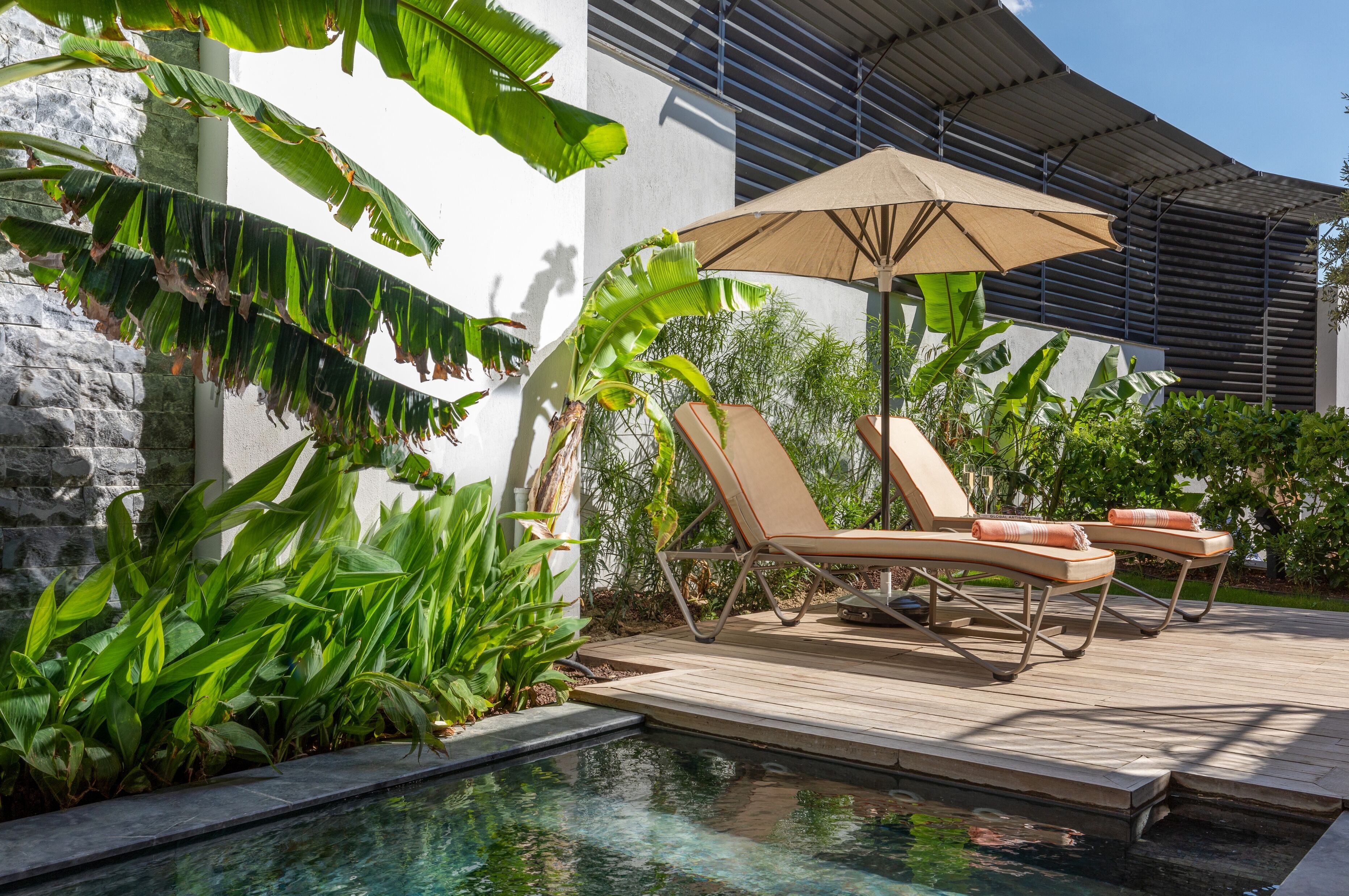 A poolside area with lounge chairs and an umbrella, surrounded by lush greenery and a modern building.