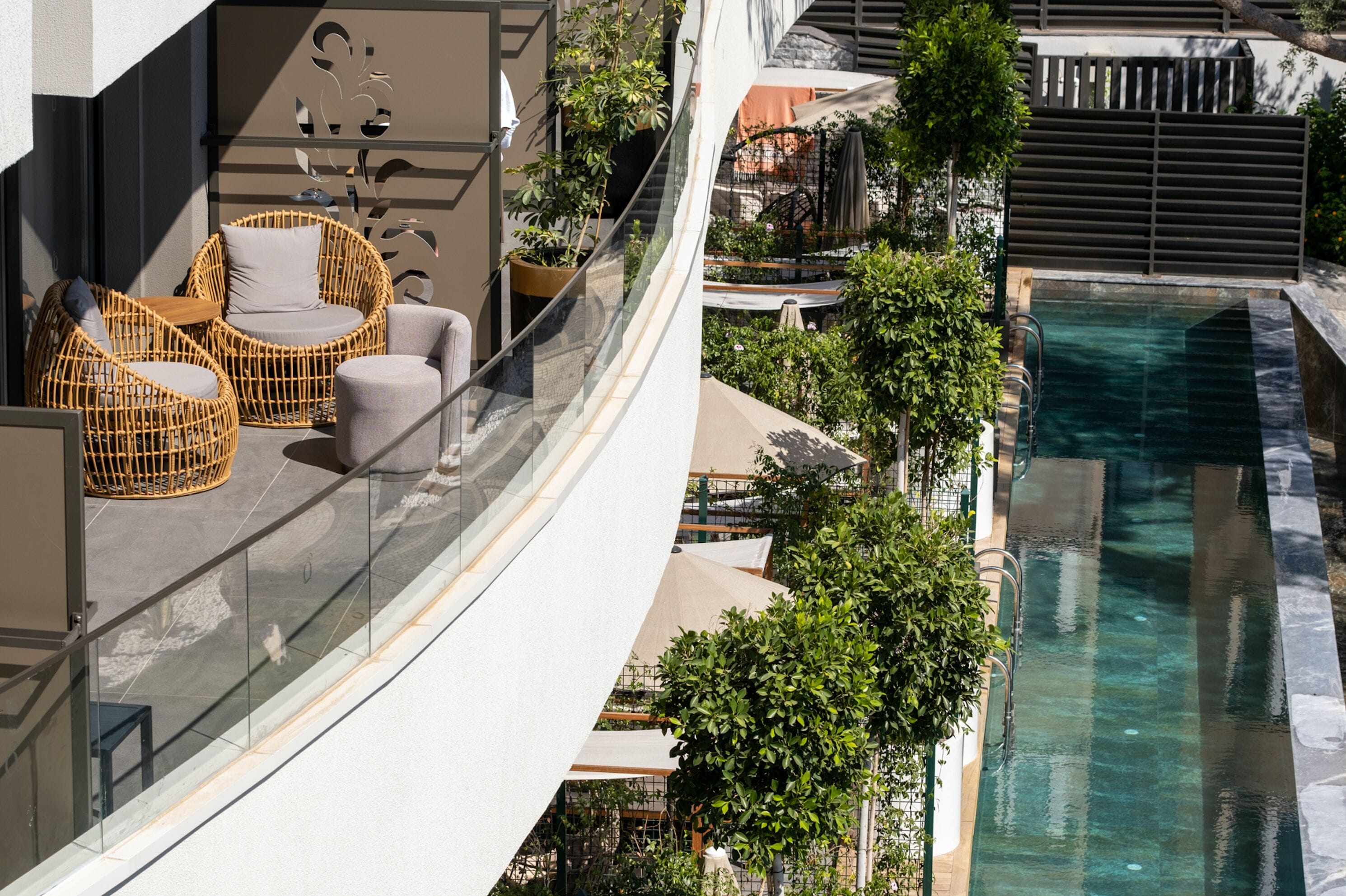 A balcony with wicker furniture, a glass railing, and a pool area with a view of the surrounding greenery.