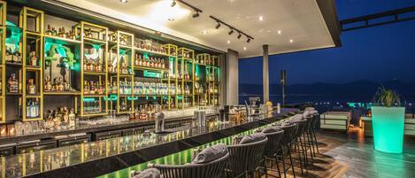 Uma área de bar com balcão extenso, várias prateleiras de garrafas e banquetas.