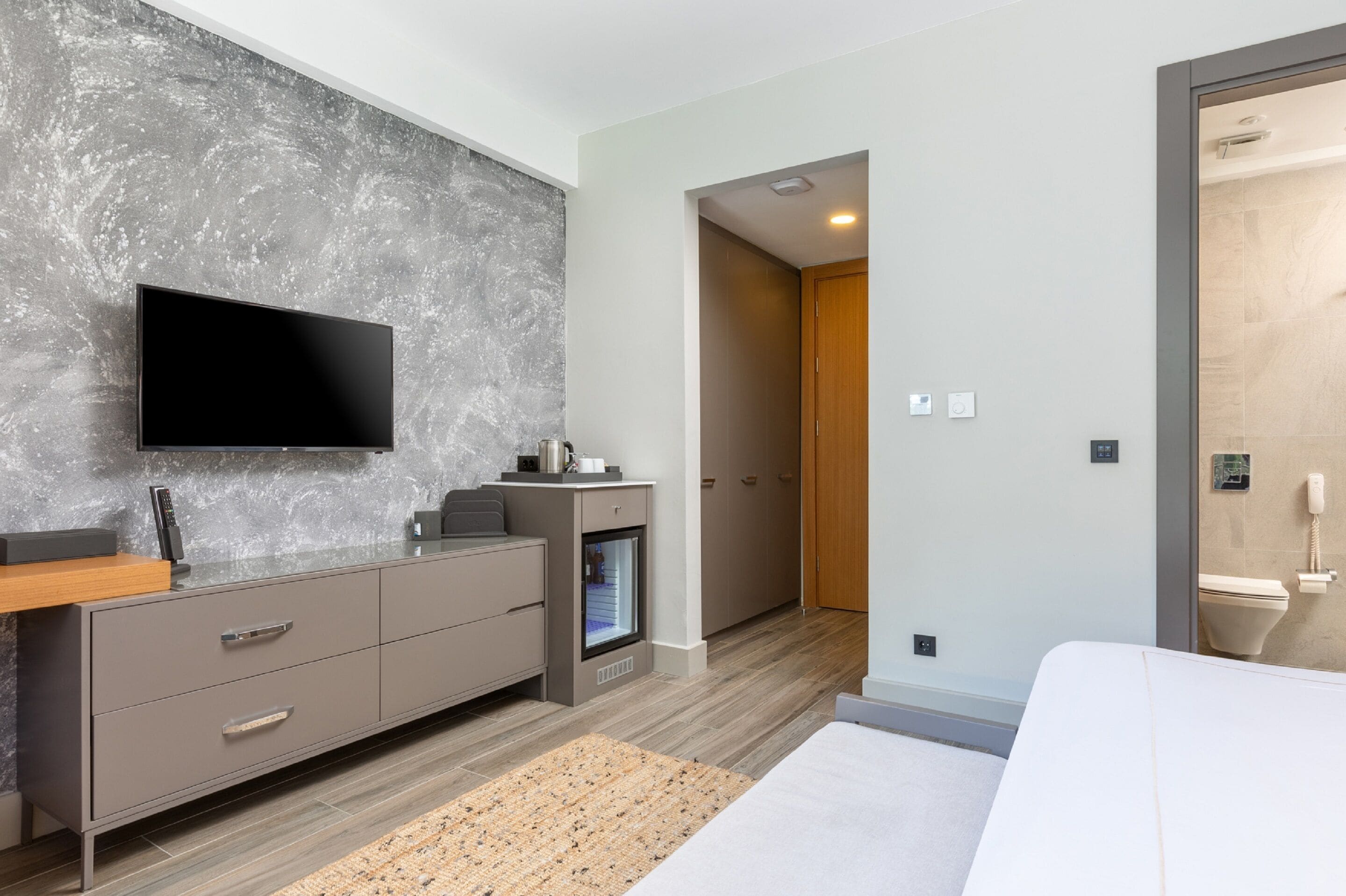 Ein modernes Hotelzimmer mit Flachbildfernseher an einer strukturierten Wand, einem eingebauten Minikühlschrank, einem Nachttisch aus Holz, einem Bett mit weißer Bettwäsche und einer Holztür zum Badezimmer.