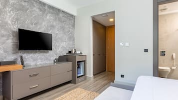 Ein modernes Hotelzimmer mit Flachbildfernseher an einer strukturierten Wand, einem eingebauten Minikühlschrank, einem Nachttisch aus Holz, einem Bett mit weißer Bettwäsche und einer Holztür zum Badezimmer.