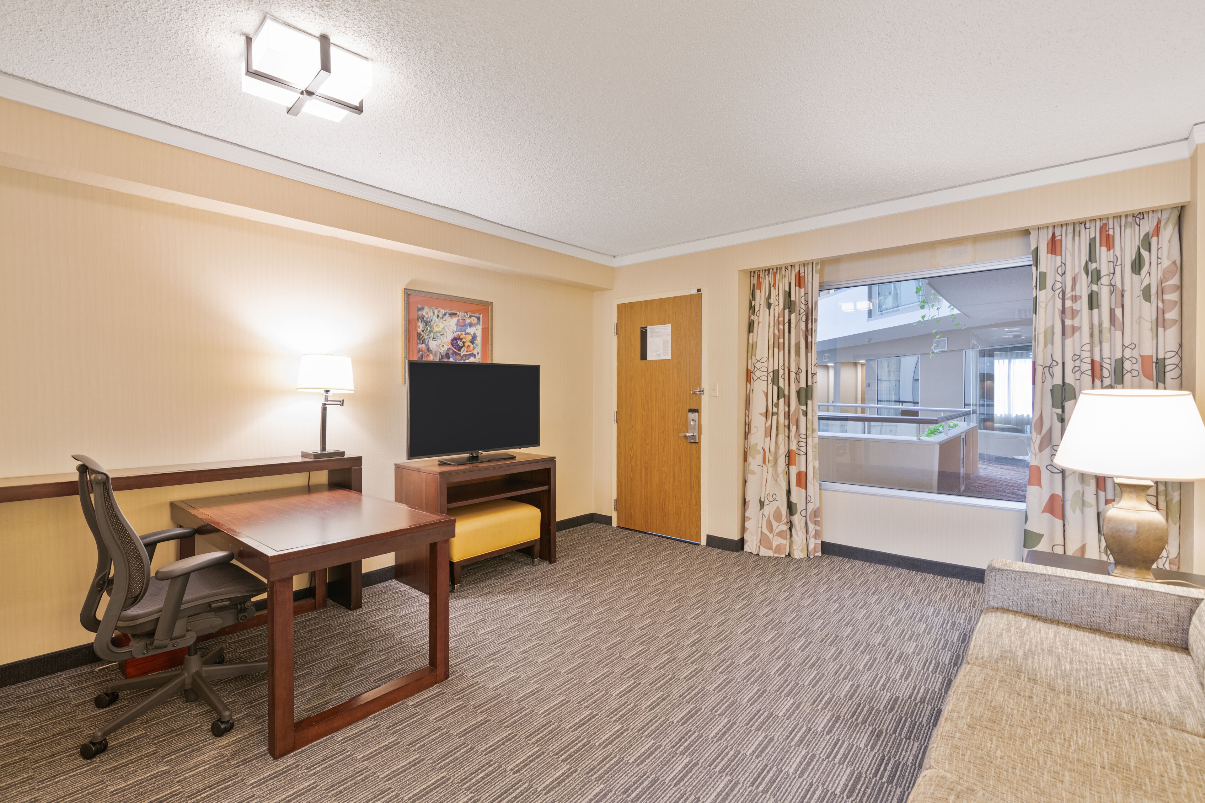 A hotel room with a wooden desk, a television, a sofa, and a window with curtains.