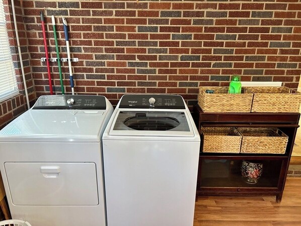 Une buanderie équipée de deux machines à laver et deux sèche-linge blancs, un mur en briques et des paniers en osier posés sur une étagère en bois.