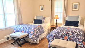 Ein Schlafzimmer mit zwei Betten, jedes mit einem gemusterten Bettbezug und passenden Kissen. Zwischen den Betten steht ein kleiner Tisch mit einer Lampe, und am Fußende jedes Bettes eine Bank.