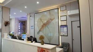 A reception area with a white counter, a large framed artwork, a small plant, and a chair. - GreenTree Inn Express Hotel (Haikou Haixiu Middle Road Bangbaihui City Branch) (Haikou)