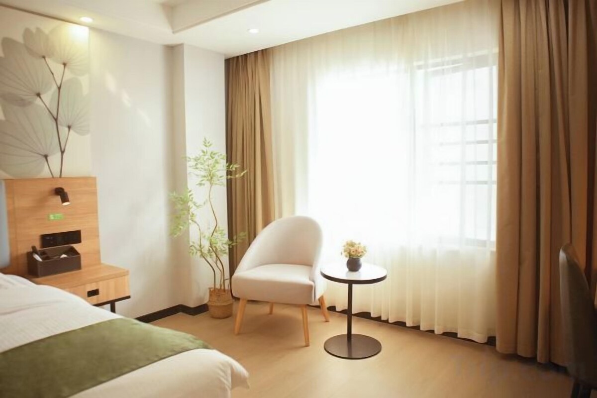 Habitación de hotel con una cama, una silla, una mesita con un jarrón, una planta y una ventana con cortinas.