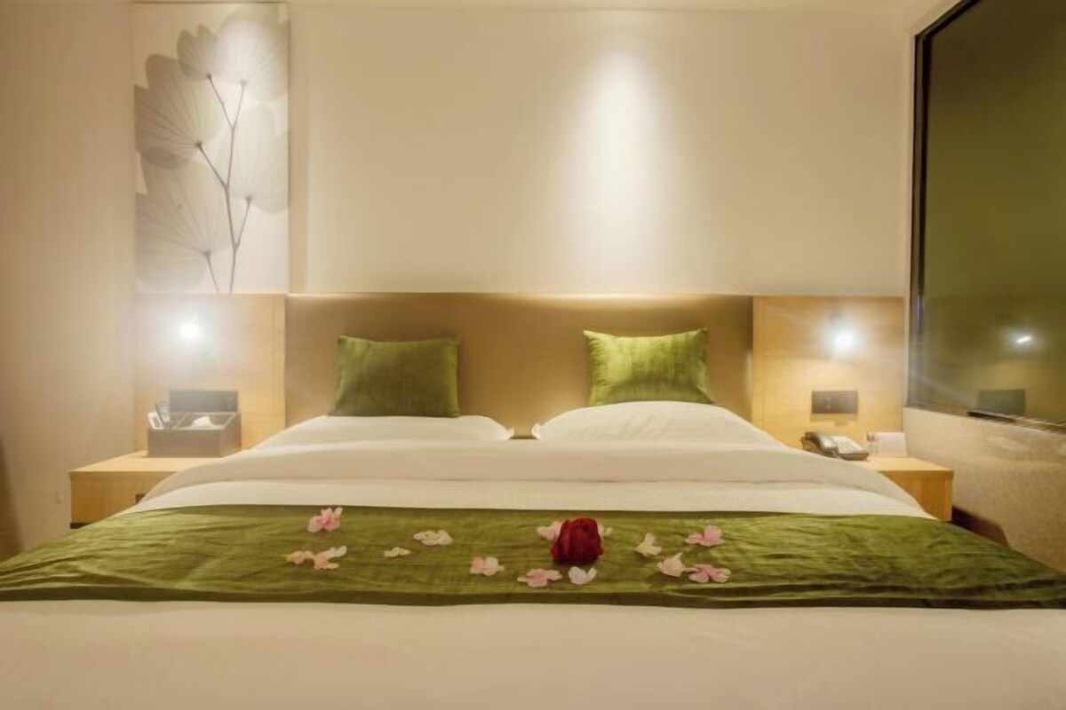 A hotel room with a bed, green pillows, a bedside table with a phone, and a wall-mounted artwork.