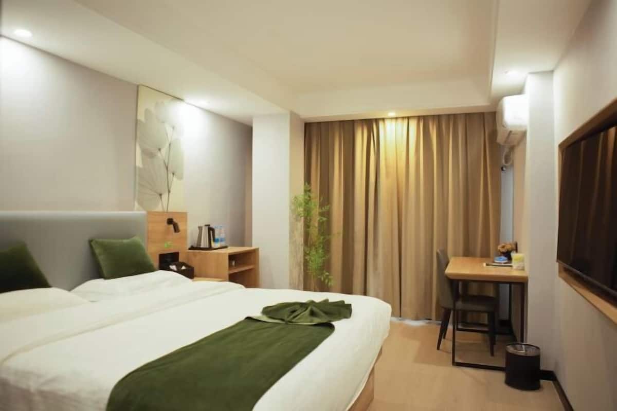 Hotellihuoneessa on suuri sänky, työpöytä ja tuoli. Siellä on myös kasvi, roskakori ja ikkuna, jossa on verhot.