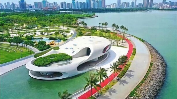 A modern building with a unique curved design, situated by a waterfront with a park and a road. - GreenTree Inn Express Hotel (Haikou Haixiu Middle Road Bangbaihui City Branch) (Haikou)