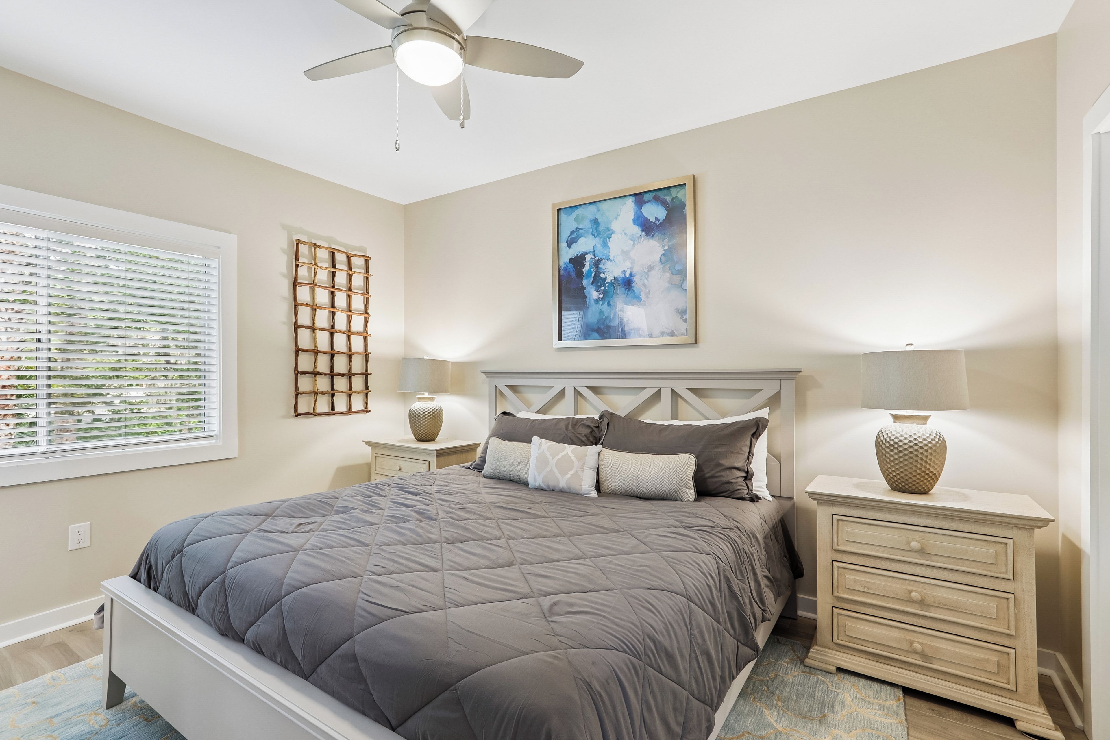 A bedroom with a bed, a ceiling fan, a window with blinds, a wall-mounted abstract painting, and bedside lamps.