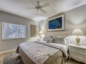 Une chambre à coucher comprenant un lit, deux tables de chevet, un ventilateur de plafond, une fenêtre avec des stores, une œuvre d’art accrochée au mur et un tapis.