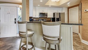 Una cocina con una isla central, dos taburetes de bar, microondas, horno y refrigerador.