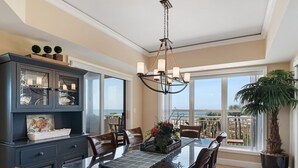 A dining area with a glass table, wooden chairs, a dark blue cabinet with glass doors, and a view of the ocean through large windows. - Direct Ocean Front Villa w/ Large Balcony (Hilton Head Island)