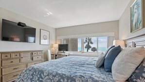 Un dormitorio con una cama grande, un televisor de pantalla plana, un escritorio con lámpara y vistas a palmeras.