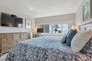 A bedroom with a large bed, a flat-screen TV, a desk with a lamp, and a view of palm trees.