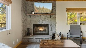 Une cheminée en pierre avec un téléviseur au-dessus, un plafond en bois et une vue sur les arbres à l’extérieur.