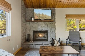 Une cheminée en pierre avec un téléviseur au-dessus, un plafond en bois et une vue sur les arbres à l’extérieur.