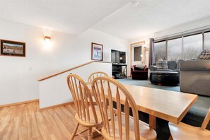 A dining area with a wooden table and chairs, a television, and a sofa in a well-lit room with large windows.