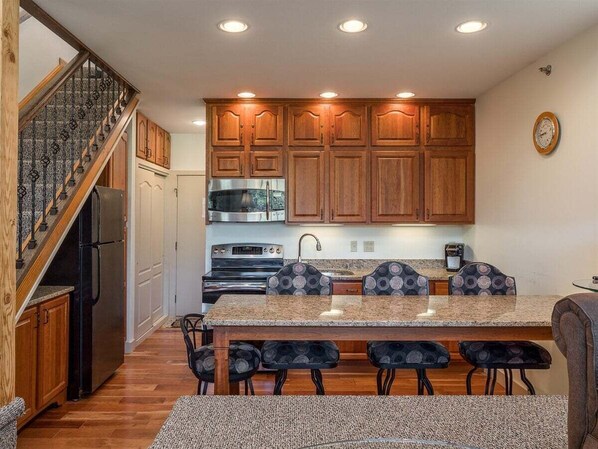 A kitchen with wooden cabinets, a stainless steel refrigerator, a built-in oven, a granite countertop, and a dining area with four chairs.