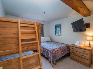 Ein Schlafzimmer mit einem Etagenbett, einem Bett mit gemusterten Bettwaren, einem Nachttisch mit Lampe sowie einem an der Wand montierten Fernseher.