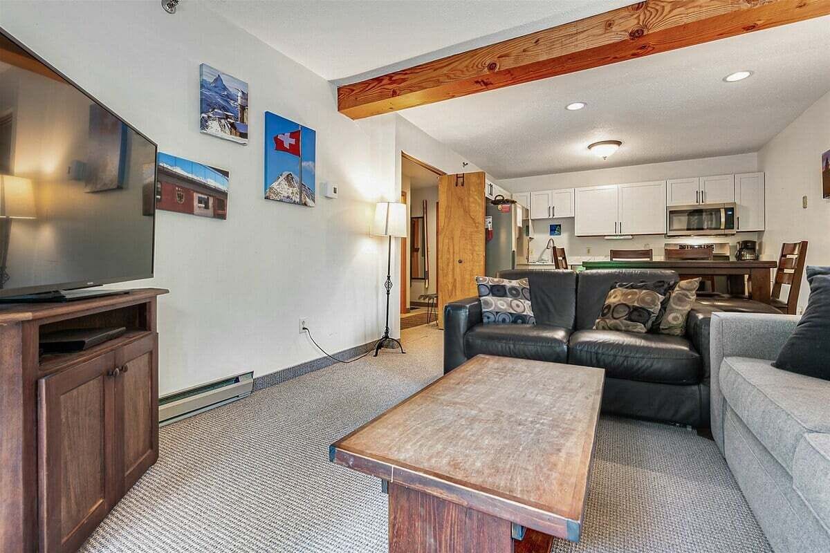 A living room with a TV, sofa, and coffee table. There is a kitchen in the background with cabinets and a microwave.