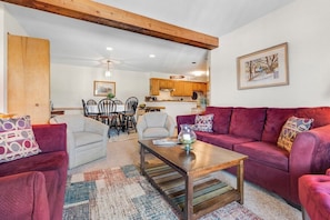 Ein Wohnzimmer mit einem roten Sofa, einem weißen Sofa und einem Couchtisch aus Holz. Im Hintergrund ist eine Küche mit Holzschränken und einer Pendelleuchte zu sehen.