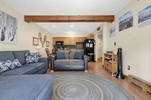 A living room with a sofa, a coffee table, and a rug. There is a kitchen in the background with wooden cabinets and a refrigerator.