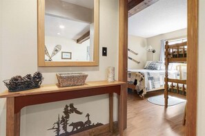 A room with a bed, a wooden console table with a basket and a decorative item, a mirror, and a bunk bed in the background.