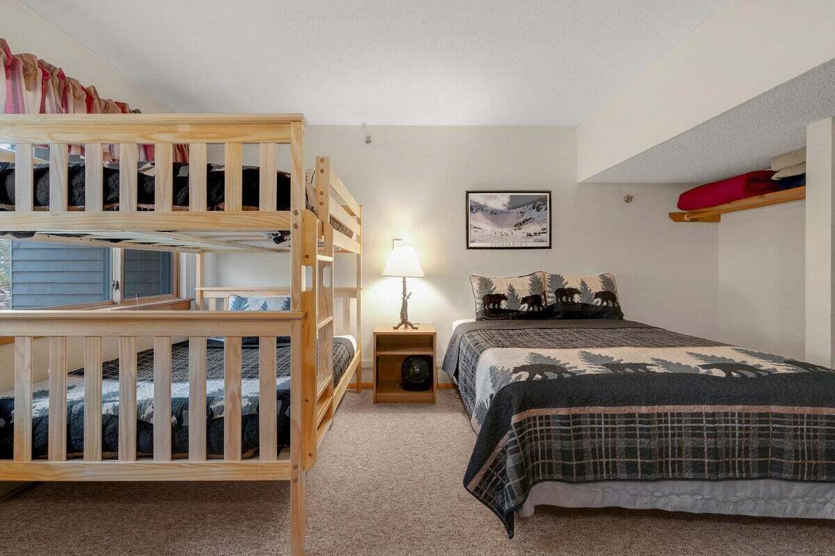 A room with bunk beds, a bedside table with a lamp, a bed with a forest-themed bedspread, and a wall-mounted shelf with a red cushion.