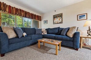 A living room with a blue sectional sofa, a coffee table with various items, a lamp, and a poster on the wall.