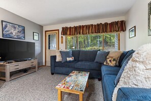 A living room with a sectional sofa, a flat-screen TV on a wooden stand, a coffee table with stickers, and a window with curtains.